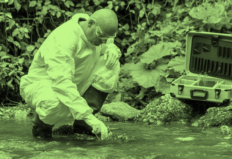 Moody employee man working to obtain water samples in blue running stream surrounded by green leaves in environment